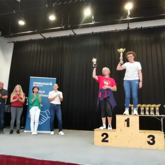 Eine Gruppe von Menschen steht auf einer Bühne, wobei drei Personen Pokale halten. Die Bühne ist mit einem Podest, Lichtern und einer Kulisse geschmückt. Andere Menschen sind anwesend, einige klatschen.