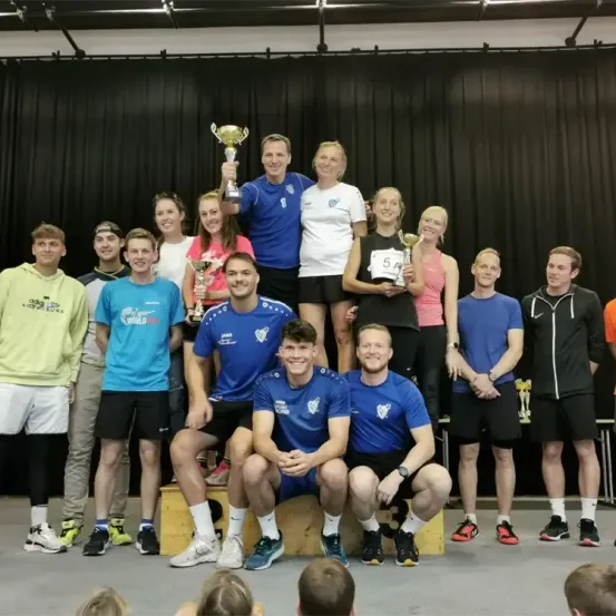 Eine Gruppe von Menschen posiert für ein Foto auf einer Bühne, einige halten Pokale. Sie tragen verschiedene Sportbekleidung und Schuhe. Der Mann in der Mitte lächelt und hält einen Pokal. Die Person vorne sitzt auf einer Holzkiste. Hinter ihnen befindet sich ein schwarzer Vorhang.