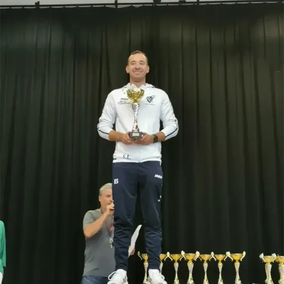 Ein Mann steht auf der Bühne, hält einen Pokal und lächelt. Ein Mann hinter ihm spricht. Mehrere Trophäen sind vor ihnen aufgestellt.