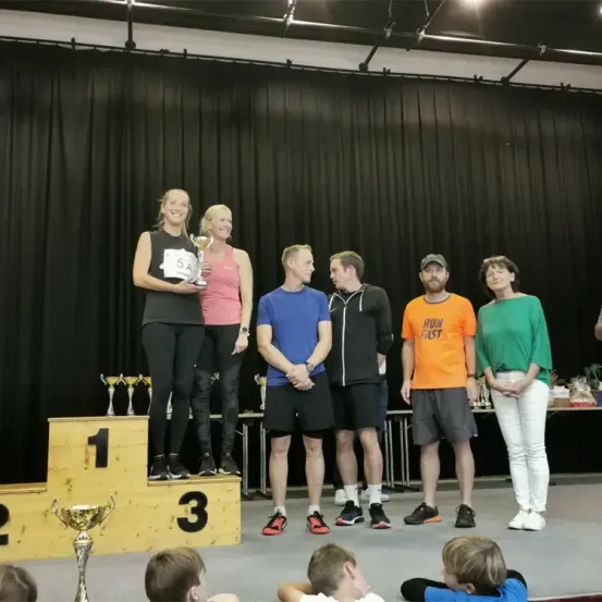 Die Gewinner stehen auf dem Podest und halten eine Trophäe in den Händen, während andere Teilnehmer dahinter stehen. Die Bühne ist mit schwarzen Vorhängen und Trophäen dekoriert. Kinder sind bei der Veranstaltung anwesend.