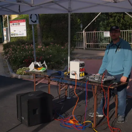 Ein Mann steht unter einem Vordach und benutzt einen DJ-Mixer auf einem Tisch mit Kabeln und einem Lautsprecher. Hinter ihm steht ein Schild und ein Tor.