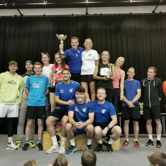 Eine Gruppe von Menschen posiert für ein Foto auf der Bühne, hält Pokale in den Händen, zwei Männer sitzen vorne in blauen Trikots. Hinter ihnen stehen andere Menschen, einige stehend und einige sitzend. Ein schwarzer Vorhang ist hinter ihnen.