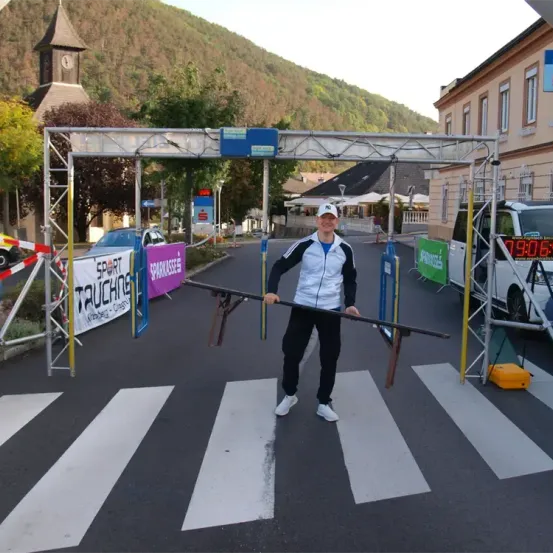 Ein Mann steht an einer Fußgängerschranke, trägt eine Mütze und Turnschuhe. Hinter ihm steht ein Gebäude mit einem Glockenturm, und Banner hängen an der Seite. Ein weißer Van ist in der Nähe geparkt.
