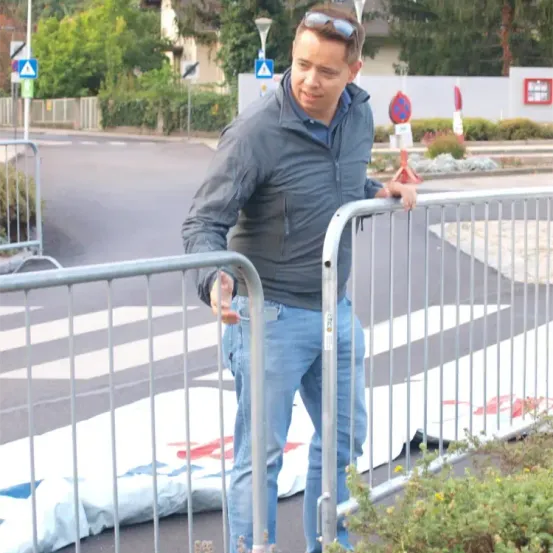 Ein Mann steht auf einem Bürgersteig und hält sich an einer Metallbarriere fest. Er trägt eine Sonnenbrille auf dem Kopf, ein blaues Hemd und hellblaue Jeans. Hinter ihm ist ein Zebrastreifen mit weißen Streifen markiert.