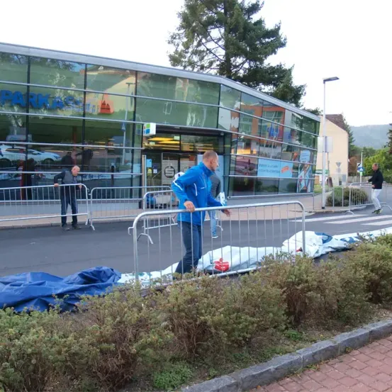 Ein Mann in einer blauen Jacke geht neben einem Gebäude mit Glaswänden. Zwei Personen stehen in der Nähe einer Metallabsperrung. Hinter der Absperrung befinden sich Büsche und ein Auto ist vor dem Gebäude geparkt.