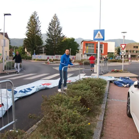 Ein Mann steht an einer Absperrung auf einer Straße, möglicherweise in Vorbereitung auf eine Veranstaltung. In der Nähe gehen Menschen über die Straße, und im Hintergrund befinden sich Gebäude und Bäume.