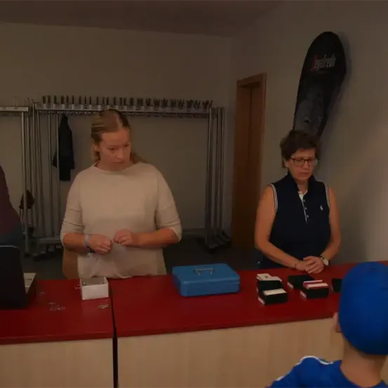 Zwei Frauen stehen hinter einem Tresen, eine bereitet etwas vor, während die andere zuschaut. Sie haben Boxen und eine blaue Tasche vor sich. Ein Kind sitzt vor ihnen.