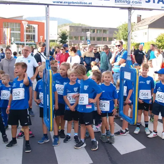 Kinder in blauen Trikots mit Nummern stehen am Start eines Rennens, mit Zuschauern und Gebäuden im Hintergrund.
