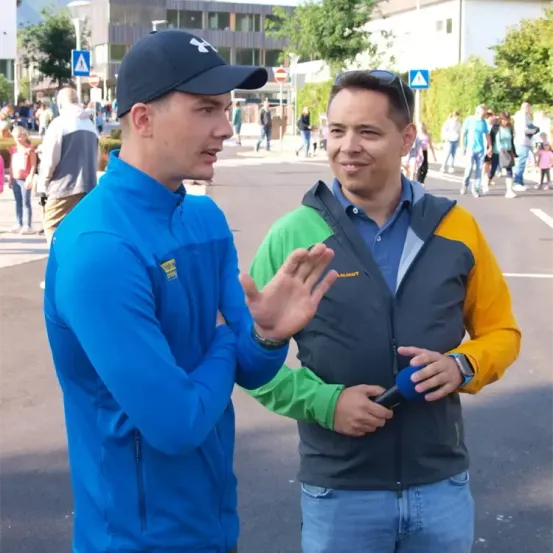 Zwei Männer stehen auf der Straße, einer hält ein Mikrofon und führt scheinbar ein Interview. Der Mann in Blau und Schwarz spricht, während der andere aufmerksam zuhört. Hinter ihnen schlendert eine Menschenmenge die Straße entlang.