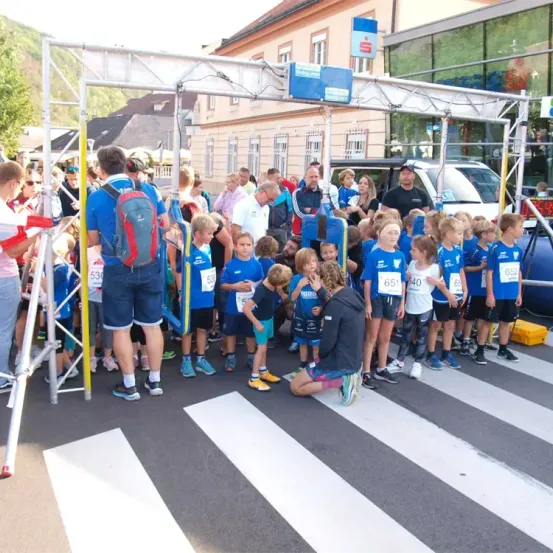 Eine Gruppe von Kindern in blauen Shirts mit Nummern steht an der Startlinie eines Rennens, begleitet von Erwachsenen. Die Szene umfasst eine Metallstruktur und ein Gebäude im Hintergrund.