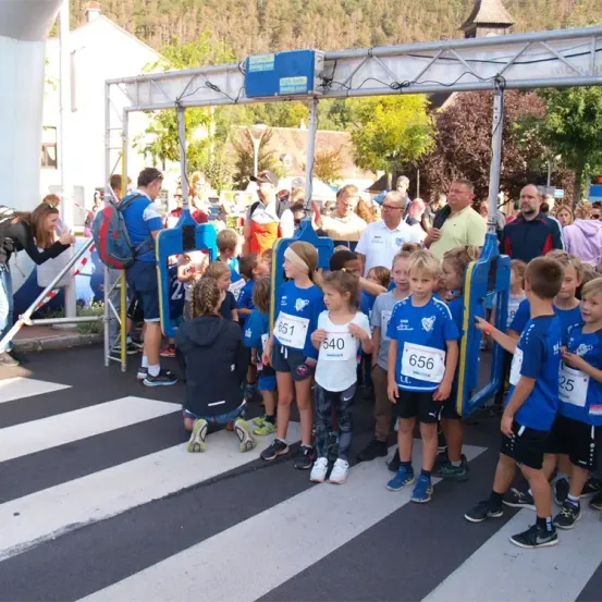 Kinder in blauen Trikots mit Nummern stehen am Start eines Rennens, umgeben von Erwachsenen und Zuschauern. Hinter ihnen befindet sich eine Metallkonstruktion.