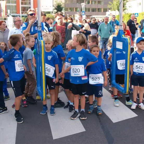 Eine Gruppe von Kindern in blauen Shirts mit nummerierten Startnummern steht in einer Reihe an einem Zebrastreifen. Zuschauer sind hinter ihnen versammelt, einige halten Kameras. Im Hintergrund befinden sich Gebäude und Bäume.