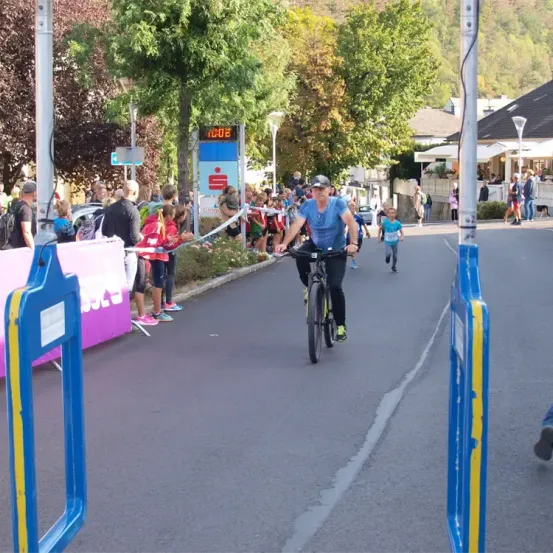 Ein Radfahrer fährt durch eine Straße mit Zuschauern. Eine Digitaluhr zeigt 11:02. Ein junger Läufer folgt. Bäume und Häuser säumen die Straße.