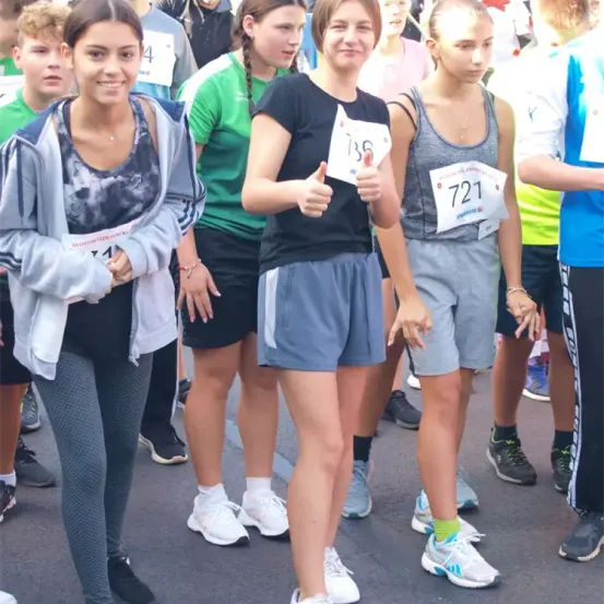 Mehrere junge Frauen stehen in einer Reihe, wahrscheinlich bei einem Marathon. Eine gibt Daumen hoch, während andere nummerierte Startnummern tragen. Die Frau rechts trägt ein Tanktop und Shorts.