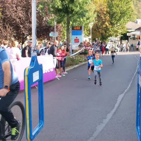 Ein Marathon mit Läufern auf einer asphaltierten Straße, Zuschauern, einer digitalen Uhr und Bäumen. Die nummerierten Läufer 526 und 567 führen.