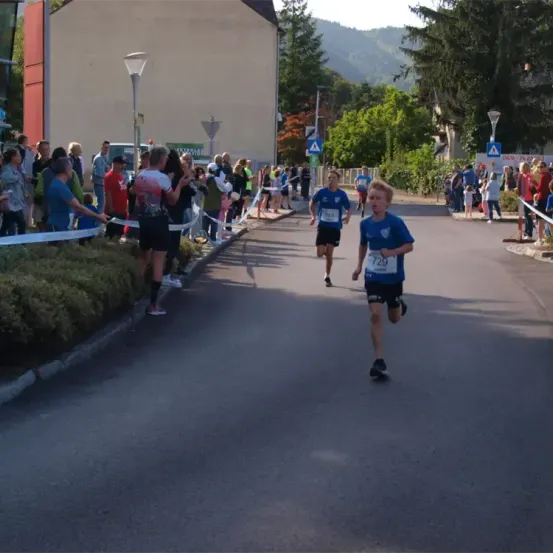 Drei Läufer nehmen an einem Marathon auf einer Straße teil, während Zuschauer sie anfeuern. Bäume und Gebäude säumen die Straße.