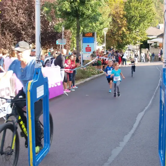 Läufer nehmen an einem Marathon auf einer Straße teil, mit Zuschauern. Eine digitale Uhr zeigt 10:08. Ein Mann auf einem Fahrrad beobachtet die Läufer.