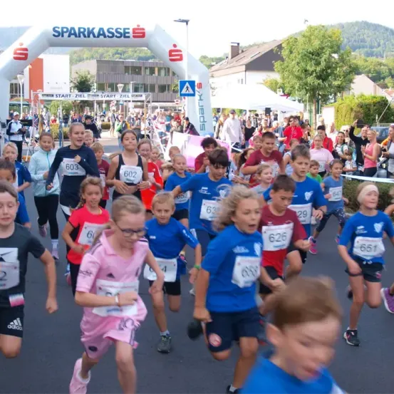 Eine große Gruppe von Kindern und Erwachsenen nimmt an einem Marathon teil. Die Kinder tragen nummerierte Trikots und laufen durch einen Parcours, der durch einen weißen Bogen gekennzeichnet ist, auf dem 'Sparkasse' steht. Zuschauer stehen an den Seiten und feuern sie an.