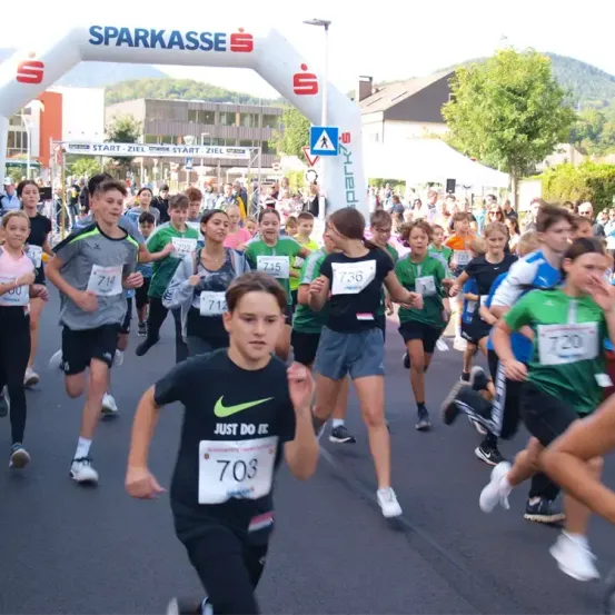 Eine Gruppe junger Leute läuft ein Rennen unter einem Torbogen mit den Worten 'Sparkasse'. Sie tragen Nummern und Turnschuhe. Im Hintergrund sind Zuschauer, Bäume und Gebäude zu sehen.