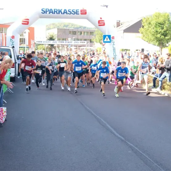 Läufer nehmen an einem Marathon teil und laufen unter einem weißen Bogen mit der Aufschrift 'Sparkasse' hindurch. Zuschauer säumen die Straße und feuern die Teilnehmer an.