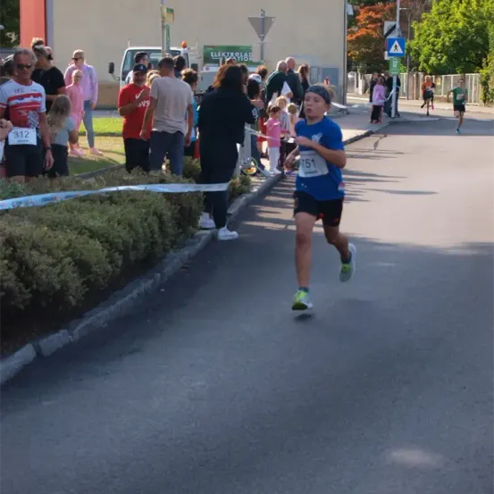 Ein Läufer mit der Nummer 51 läuft einen Marathon, während mehrere Zuschauer zuschauen. Es gibt Büsche und einen Lastwagen in der Umgebung.