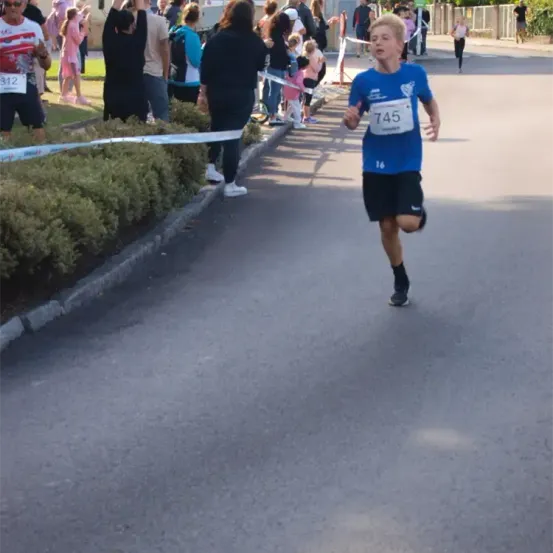 Ein junger Junge in einem blauen Shirt und schwarzen Shorts läuft in einem Rennen. Zuschauer säumen die Straße und schauen dem Rennen zu.