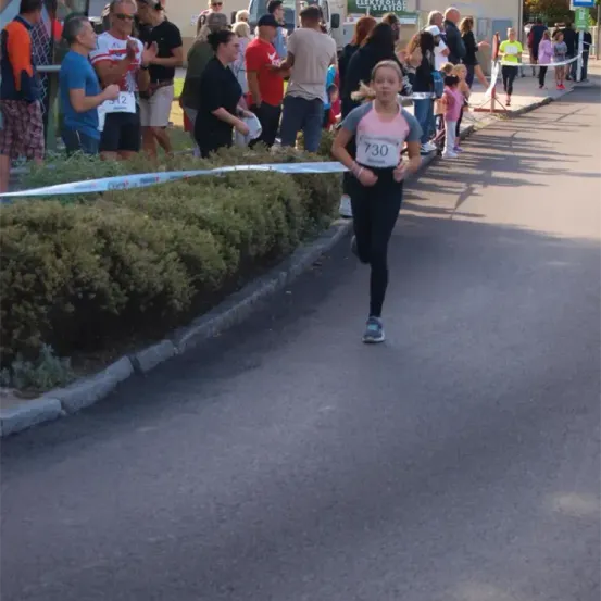 Ein junges Mädchen läuft in einem Rennen auf einer Straße mit der Nummer 730 auf ihrem Shirt, mit Zuschauern, die an der Seite zuschauen.