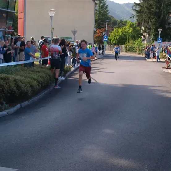 Ein Läufer in einem blauen Shirt und roten Shorts führt das Rennen an, gefolgt von mehreren anderen. Zuschauer säumen die Straße und feuern sie an. Bäume und Straßenlaternen sind am Straßenrand sichtbar.