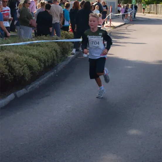 Ein Junge läuft einen Marathon. Er trägt die Startnummer 704. Zuschauer beobachten den Marathon am Straßenrand.