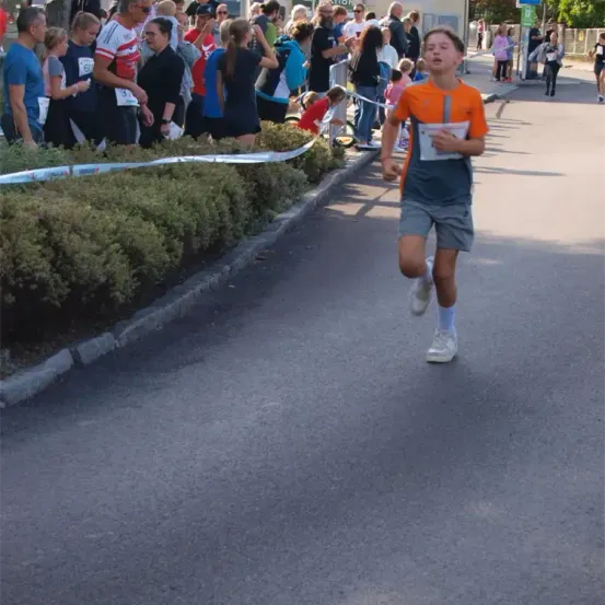 Ein junger Junge läuft in einem Rennen, während Zuschauer am Straßenrand stehen. Einige tragen Nummernschilder, während andere hinter Barrieren zusehen.