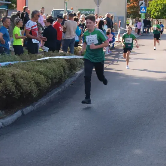 Läufer nehmen an einem Rennen auf einer Straße teil, an der eine Menge Menschen auf der Tribüne jubelt. Der Führende trägt ein grünes Shirt mit der Nummer 722.