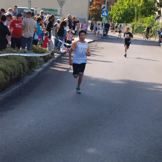 Ein junger Junge läuft in einem Rennen und trägt die Nummer 731 auf seinem Shirt. Zuschauer beobachten von der Seite, einige stehen in der Nähe eines Zauns. Bäume und Gebäude sind im Hintergrund.