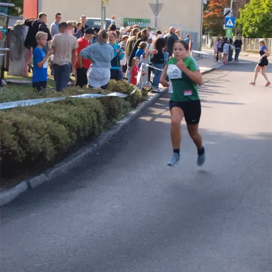 Ein junges Mädchen läuft in einem Rennen mit einer Nummer auf ihrem Shirt. Hinter ihr beobachtet eine Menschenmenge. Die Veranstaltung findet im Freien statt, und es gibt Büsche und eine Straße mit einem Gehweg.
