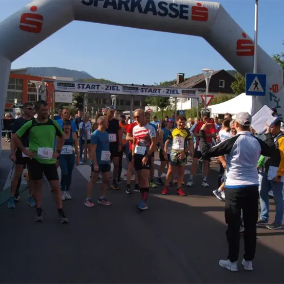 Läufer stehen am Start eines Rennens unter einem weißen Bogen mit dem Wort 'Sparkasse'. Die Leute tragen Nummern auf ihren Shirts und Shorts. Ein Mann steht vor ihnen und hält ein Papier.