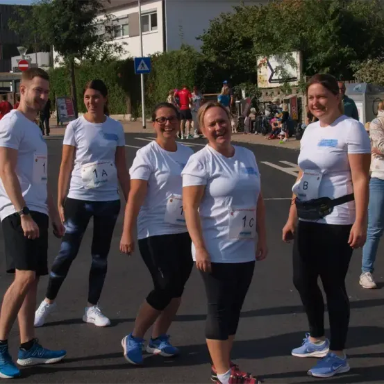 Fünf Menschen stehen auf der Straße, wahrscheinlich bei einem Marathon. Alle tragen weiße Hemden und schwarze Hosen. Sie lächeln und scheinen für ein Foto zu posieren. Hinter ihnen stehen mehrere Menschen auf dem Bürgersteig, und einige von ihnen gehen. In der Ferne gibt es mehrere Bäume, ein Gebäude und ein Schild.