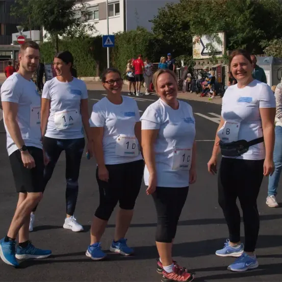 Fünf Personen in weißen T-Shirts und Laufschuhen stehen in einer Reihe, möglicherweise bei einem Marathon. Sie tragen nummerierte Startnummern. Dahinter sind verschwommene Figuren anderer Teilnehmer und Zuschauer zu sehen.