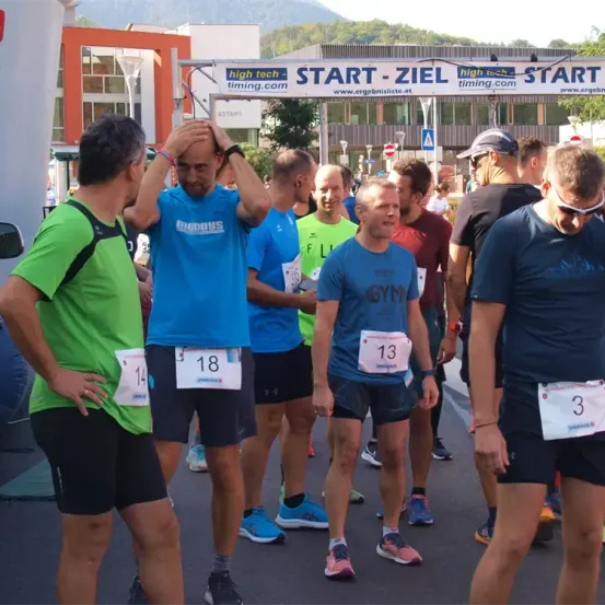 Eine Gruppe von Läufern steht an der Startlinie eines Rennens, jeder trägt nummerierte Startnummern. Im Hintergrund befindet sich ein Banner mit 'START - ZIEL' und einer Website-Adresse. Gebäude und Bäume sind in der Ferne zu sehen.