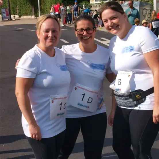 Drei Frauen in identischen weißen T-Shirts mit blauen Logos posieren für ein Foto auf einer Straße. Jede trägt eine Nummer auf ihrem Shirt: 1D, 1C und B. Sie stehen lächelnd vor einem urbanen Hintergrund.