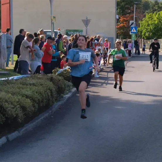 Ein Läufer mit der Nummer 711 sprintet die Straße hinunter, dicht gefolgt von einem anderen Läufer in einem grünen Shirt. Zuschauer sind entlang der Straße versammelt und beobachten das Rennen.