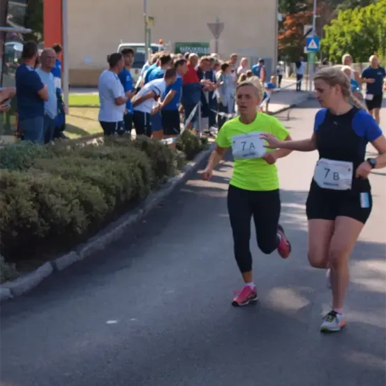 Zwei Frauen laufen in einem Marathon. Eine hat die Nummer 7A und die andere 7B. Zuschauer beobachten sie.