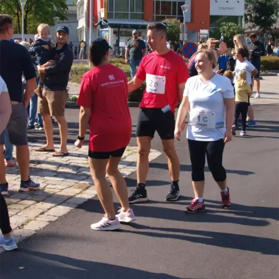 Eine Gruppe von Menschen nimmt an einem Marathon teil. Ein Mann und eine Frau unterhalten sich in der Mitte, mit den Startnummern 2D und 1C. Zuschauer, darunter ein Mann, der ein Kind hält, stehen in der Nähe.