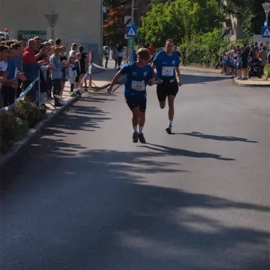 Zwei Läufer nehmen an einem Rennen teil und laufen auf einer Straße, an der Zuschauer stehen. Die Läufer tragen die Nummern 48 und 4A.