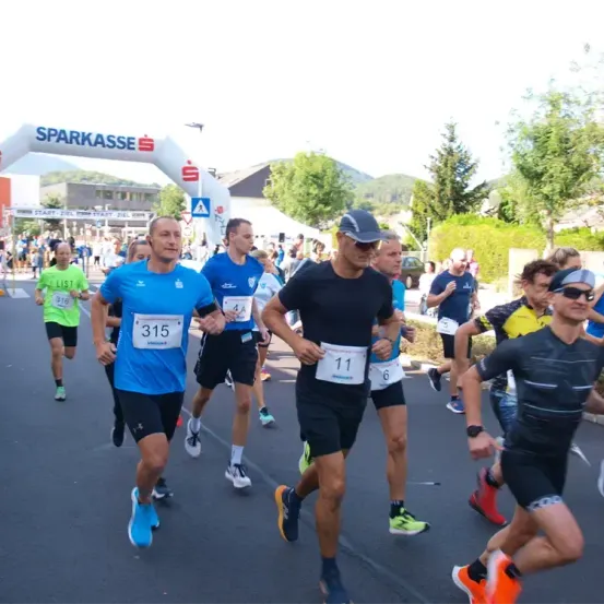 Läufer nehmen an einem Marathon teil, tragen nummerierte Trikots, ein Mann in Blau führt, ein anderer in Schwarz folgt, und Zuschauer feuern von den Seitenlinien an.