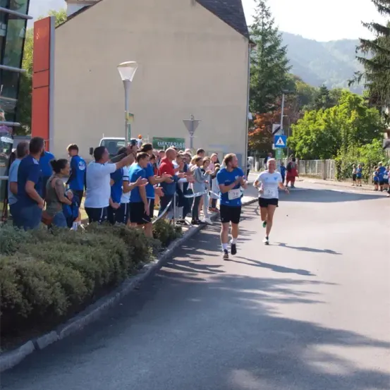 Zwei Läufer sprinten auf einer Straße, angefeuert von Zuschauern in blauen Shirts. Eine Straßenlaterne und ein Gebäude befinden sich an der Seite.
