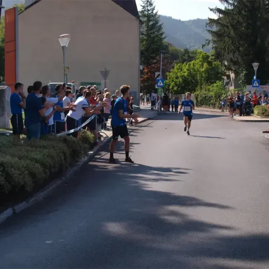 Läufer nehmen an einem Rennen auf einer Stadtstraße teil, mit Zuschauern und Straßenlaternen entlang der Strecke. Bäume und Berge bieten eine malerische Kulisse.
