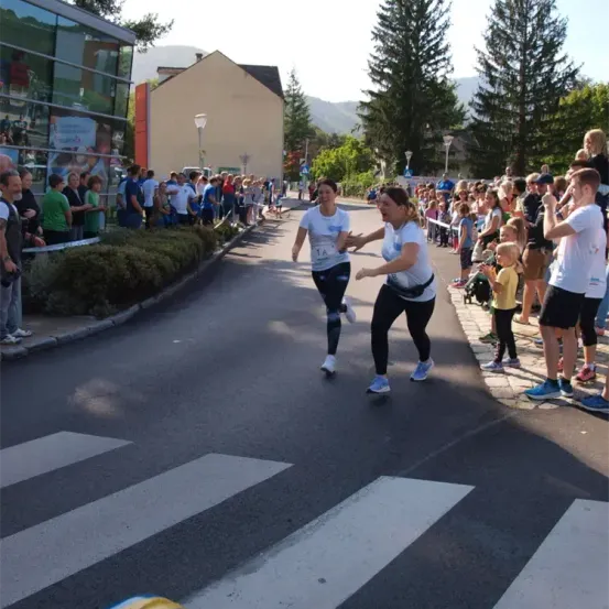 Zwei Frauen in weißen T-Shirts joggen die Straße entlang, während eine Menge zusieht. Einige klatschen und jubeln. Bäume und ein Gebäude sind im Hintergrund.