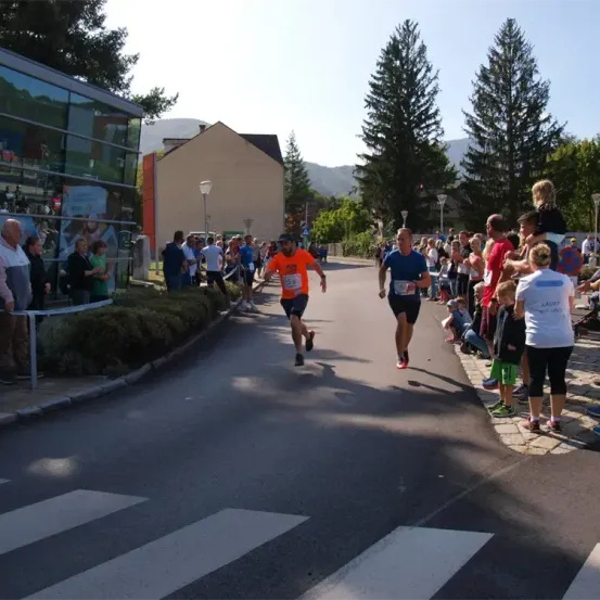 Ein Marathonlauf findet auf einer Straße statt, mit einer Menge von Zuschauern, Bäumen und Gebäuden im Hintergrund. Zwei Läufer in orangefarbenen und blauen Trikots führen.