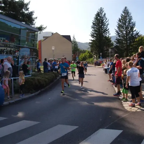 Marathonläufer sprinten eine Straße hinunter, gesäumt von Zuschauern, in der Nähe eines Gebäudes mit Glaswänden. Bäume und Berge bilden den Hintergrund.