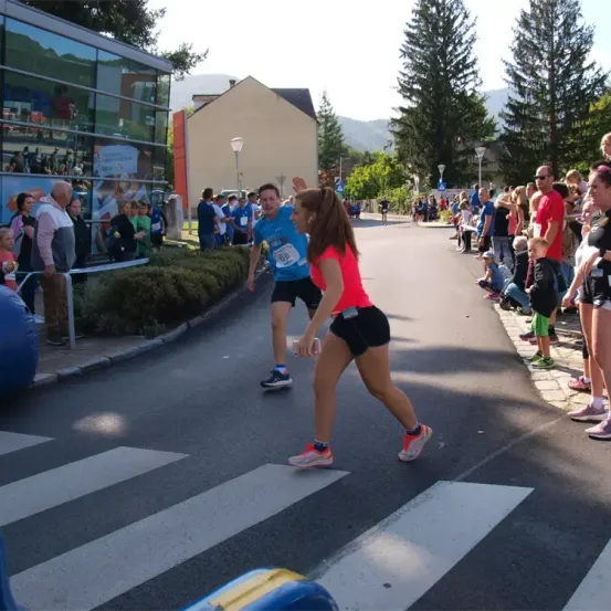 Eine Frau in einem rosa Shirt und schwarzen Shorts läuft während eines Marathons über die Straße. Hinter ihr winkt ein Mann. Zuschauer säumen den Bürgersteig.