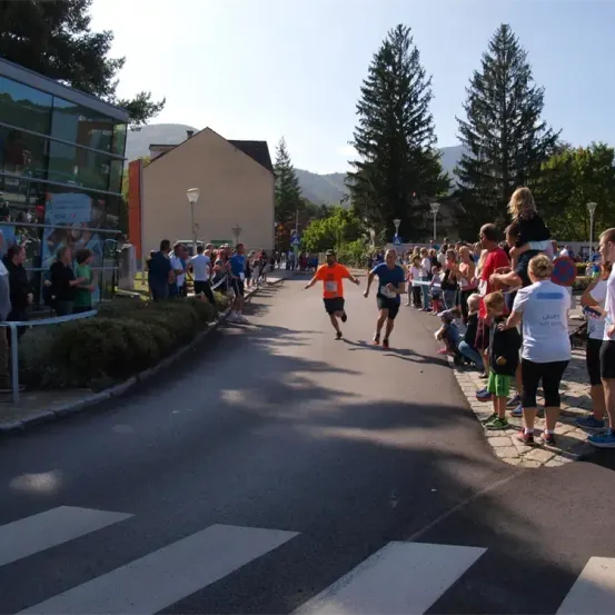 Läufer sprinten die Straße hinunter, während Zuschauer auf beiden Seiten jubeln. Im Hintergrund sind Bäume und Gebäude zu sehen.